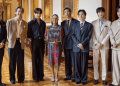 Group of eight people in formal suits posing in a grand room; a woman in a patterned dress holds a poster at center.