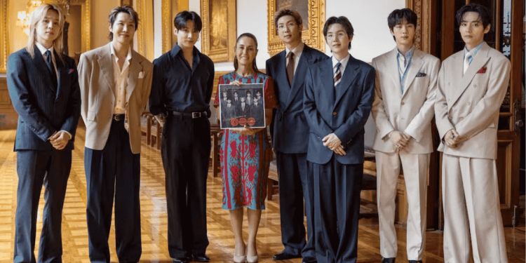 Group of eight people in formal suits posing in a grand room; a woman in a patterned dress holds a poster at center.
